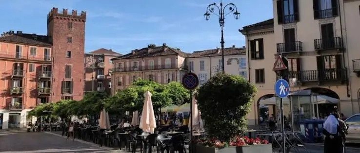 Piazza cittadina ad Asti con tavolini all’aperto, alberi verdi, lampione e palazzi storici sotto cielo azzurro.