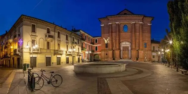 Piazza illuminata ad Alessandria con chiesa in mattoni, fontana centrale e biciclette parcheggiate.