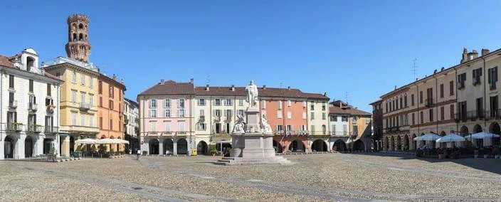 Piazza cittadina di Vercelli con fontana centrale, edifici storici colorati e cielo azzurro limpido.