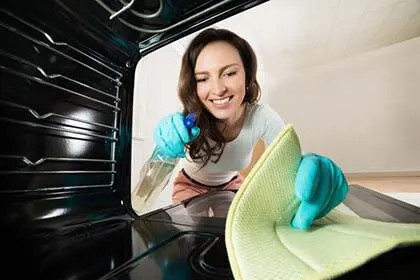 Donna sorride mentre pulisce un forno LG con guanti azzurri e panno verde, in cucina luminosa.